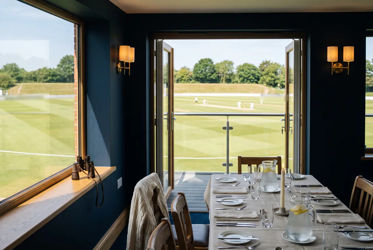 A lunch table set in a navy-walled box with brass wall lights, open doors onto a balcony above a cricket match on a summer afternoon