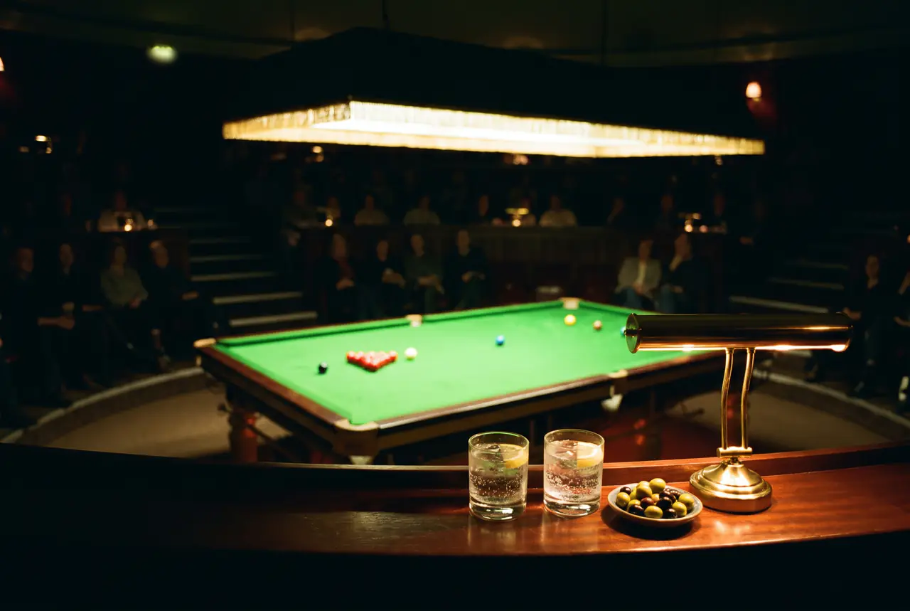 Two glasses of sparkling water, olives and a brass lamp on a ledge above a snooker table lit under its canopy, the audience in shadow